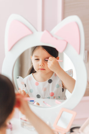 a beautiful little girl in the mirror preening. a little girl is sitting at a children's table and is wearing children's cosmetics.の写真素材