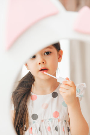a little girl looks in the mirror and paints her lips with a pink baby glitter.の写真素材