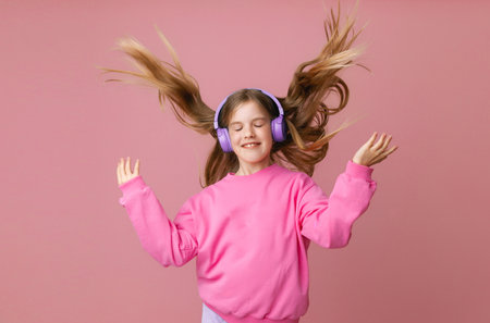 Photo of a cute funny young teenage girl in a jumper, headphones, dancing, enjoying music on a pink backgroundの写真素材