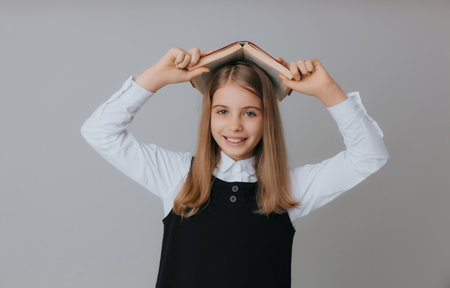 A roof over your head book. A little girl is holding a textbook on her head. Little school girl with a book on a gray background. back to school.の写真素材