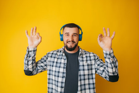 Happy caucasian guy shows a good gesture, agrees with something, claims that everything is fine, smiles broadly, wears headphones, enjoys isolated on a yellow backgroundの写真素材