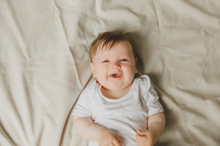 Charming blue-eyed 6-month-old baby lies in bed in a white bodysuit. View from above