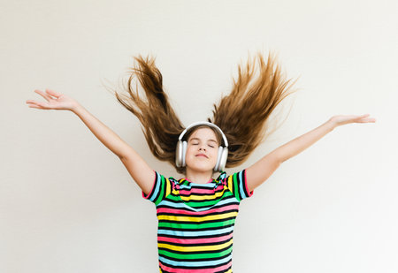 An energetic girl wearing headphones listens to music on a gray background in the studio. She wears a striped T-shirt. Long hair flies from movement.の写真素材