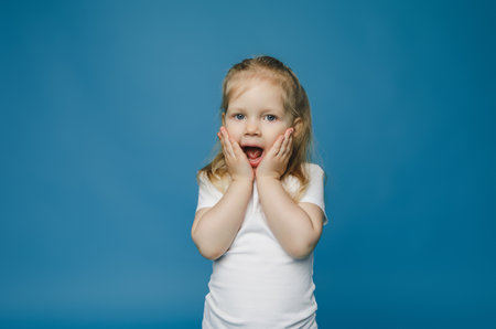 cute little girl 2-3 years old with blond hair looks at camera holds her face and is surprised.