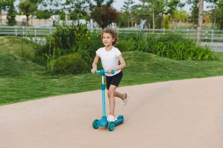 A little curly-haired girl rides a scooter in a city park.の写真素材