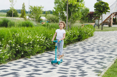A little curly-haired girl rides a scooter in a city park.の写真素材