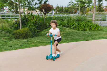 A little curly-haired girl rides a scooter in a city park.の写真素材
