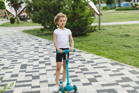 A little curly-haired girl rides a scooter in a city park.の写真素材