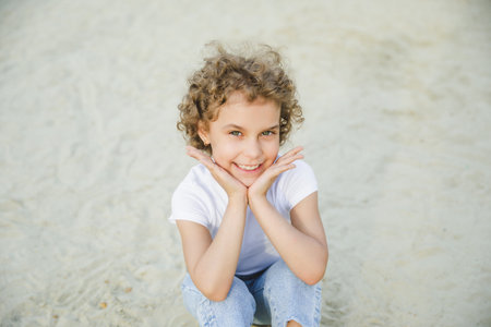 portrait of a little curly cheerful blonde girl sitting on the sandの写真素材