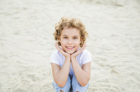portrait of a little curly cheerful blonde girl sitting on the sandの写真素材