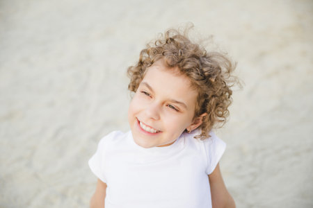 portrait of a little curly cheerful blonde girl sitting on the sandの写真素材