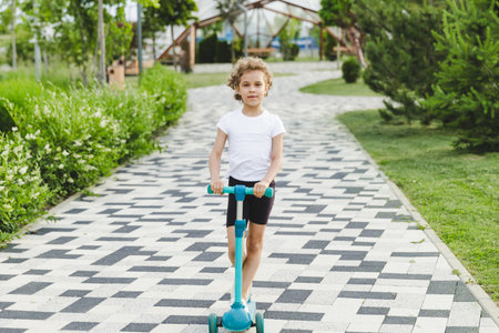A little curly-haired girl rides a scooter in a city park.の写真素材