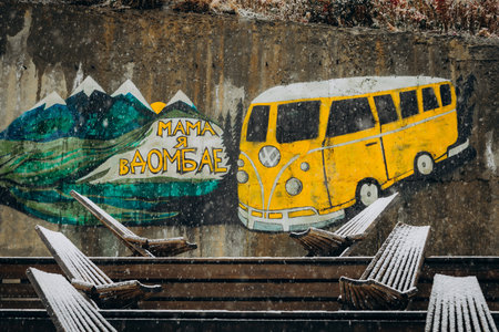 Street art mural in Dombay showing a yellow VW bus next to mountains and the phrase "Mama, I am in Dombay." Snowy weather covers the wooden chairsの写真素材