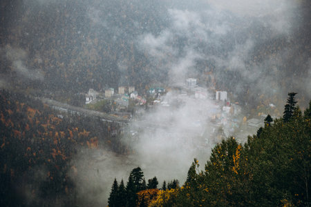 Dramatic high-angle view of the Dombay village nestled in the Caucasus valley, shrouded in fog and snow, contrasting with the surrounding autumn forestの写真素材