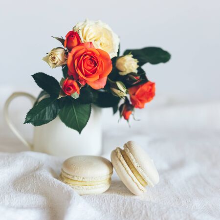 Two cream French macarons with tender cream and coral roses in a jar on white background. Close up.の写真素材