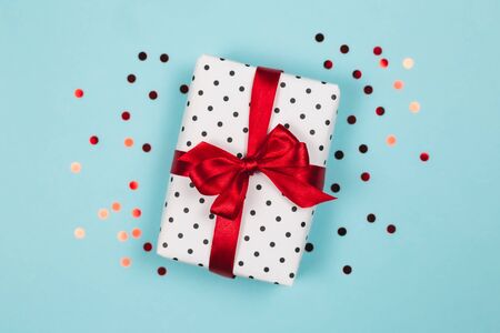 Gift box wrapped in black polka-dot white paper with red bow on blue background with confetti. Holiday concept. Flat lay.の写真素材