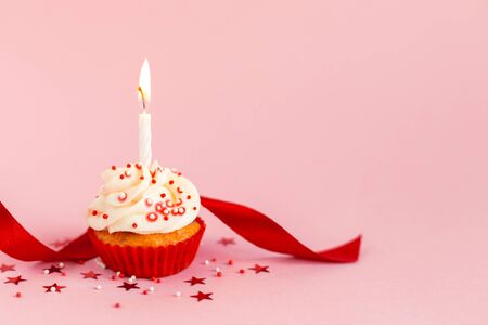 Birthday cupcake with buttercream icing, red ribbon and candle on pink pastel background. Place for text.の写真素材