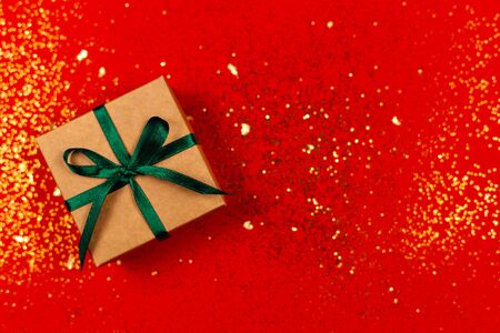 Craft gift box with green bow on red background with golden sparkles. Holiday concept. Flat lay. Place for text.の写真素材