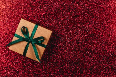 Craft gift box with green bow on dark red sparkle background. Holiday concept. Flat lay. Place for...