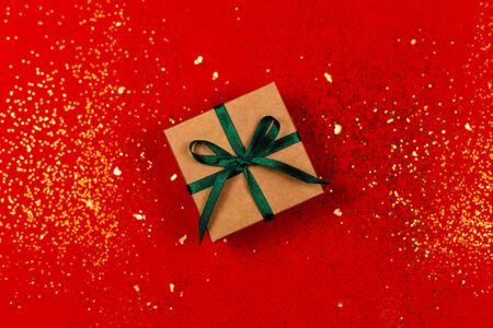 Craft gift box with green bow on red background with golden sparkles. Holiday concept. Flat lay.の写真素材