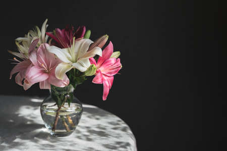 Romantic bouquet of the white and pink lilies in a glass vase on a dark background in sunlight. Place for text.の写真素材