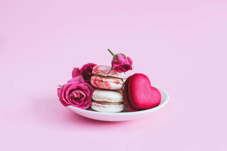Tasty french macaroons with pink roses on a white plate. Heart-shaped macaron. Pink pastel background. Place for text.の写真素材