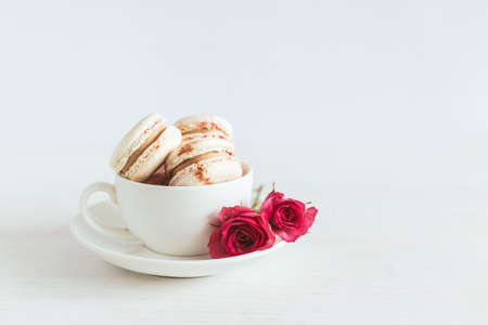 Tasty french macaroons with pink roses in a white cup. White wooden background. Place for text.の写真素材