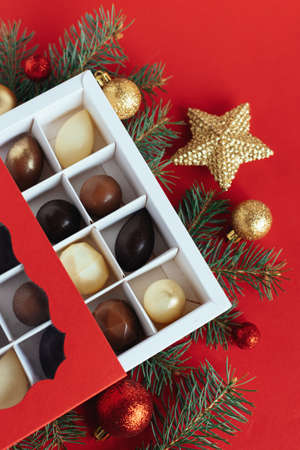 Gift box with chocolate candies, spruce branches, red and golden balls on a red background. Holiday concept. Place for text. Flat lay.の写真素材