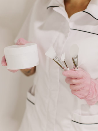 Facial clay mask in white jar and brushes in a hands of a cosmetologist. Skin care beauty product....