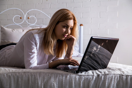 A smiling woman lying down the bed in front of her laptop with her legs raised slightlyの写真素材