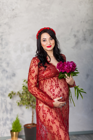 Happy pregnant woman in a red lace dress stroking her tummy against a background of huge flowers, looking to the cameraの写真素材