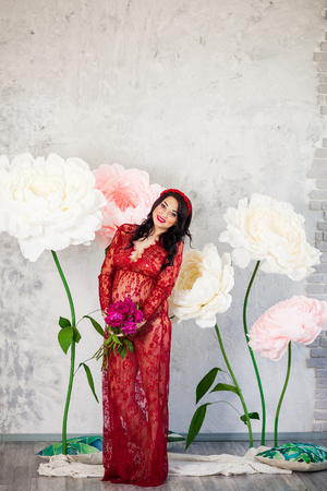Happy pregnant woman in a red lace dress stroking her tummy against a background of huge flowers, looking to the cameraの写真素材
