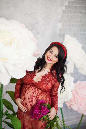 Happy pregnant woman in a red lace dress stroking her tummy against a background of huge flowers, looking to the cameraの写真素材