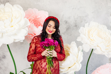 Happy pregnant woman in a red lace dress stroking her tummy against a background of huge flowers, looking at flowersの写真素材