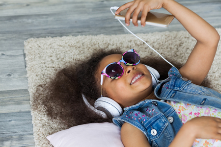 cute African girl at home, listening to music in big white headphones, Lies on the floorの写真素材
