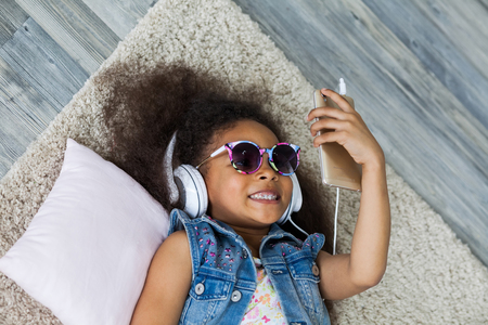 cute African girl at home, listening to music in big white headphones, Lies on the floorの写真素材