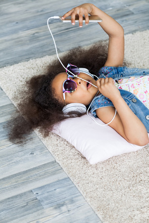 cute African girl at home, listening to music in big white headphones, Lies on the floorの写真素材