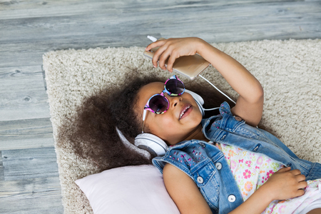 cute African girl at home, listening to music in big white headphones, Lies on the floorの写真素材