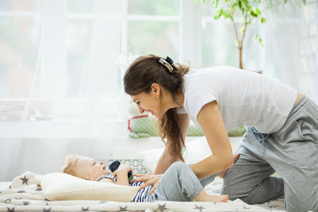 young mother with her one years old little son dressed in pajamas are relaxing and playing in the bedroomの写真素材