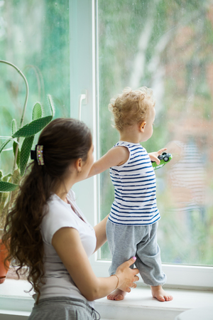 Young mother and Adorable little blond child stands near window and looking on raindrops, indoorsの写真素材