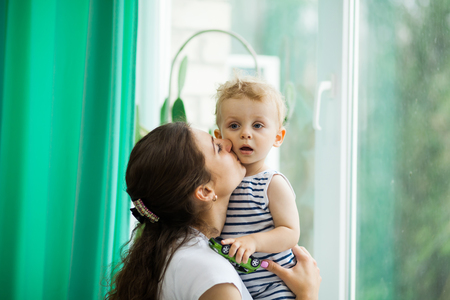 Young mother and Adorable little blond child stands near window and looking on raindrops, indoorsの写真素材