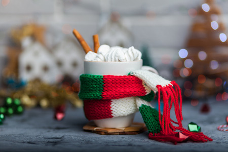 White cup of hot cocoa with mini marshmallows and cinnamon with green red white scarf against a rustic background with beautiful Christmas lights of bokeh with toysの写真素材