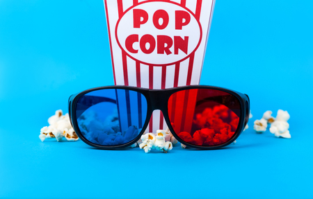 Cinema background. Film watching. Loose popcorn in striped box and 3d glasses on blue backgroundの写真素材