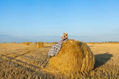 Adorable girl wearing straw hat in the village on a haystack, drink milk and eat baked goods in wheat field on warm and sunny summer evening. Cute little child in rye field on sunset.の写真素材