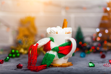 Mug Of Hot Coffee Or Tea with mini marshmallows and cinnamon with green red white scarf against a rustic background with beautiful Christmas lights of bokeh with toysの写真素材