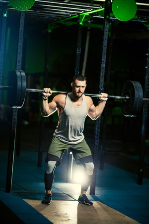 Strong motivated muscular bodybuilder man holding a heavyweight barbell on the shoulder behind the neck while crouching and doing squats exercise in the dark gym.の写真素材