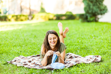 A young woman sitting in a city park on green grass and knitting a blue sweater with knitting needles on a summer day. Get Creative everywhereの写真素材