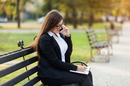 Happy businesswoman calling on mobile phone and taking notes to notepad on the street, outdoorの写真素材