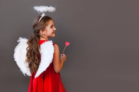 A little Cupid angel girl in a red dress with wings and halo holding red hearts in her hands, on a...