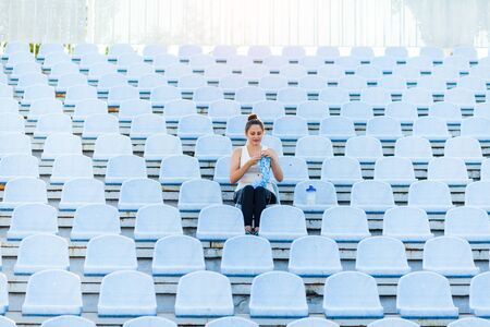 A young dark-haired woman in a sports suit at the empty stadium kniting a blue sweater. Get Creative...
